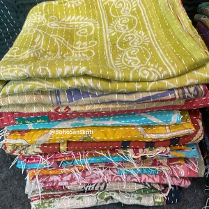 May include: A stack of colourful kantha quilts with intricate embroidery and stitching. The top quilt is a light green with white floral designs. The quilts are folded and stacked on top of each other. The text "@BohoSanskruti" is visible on the stack of quilts.
