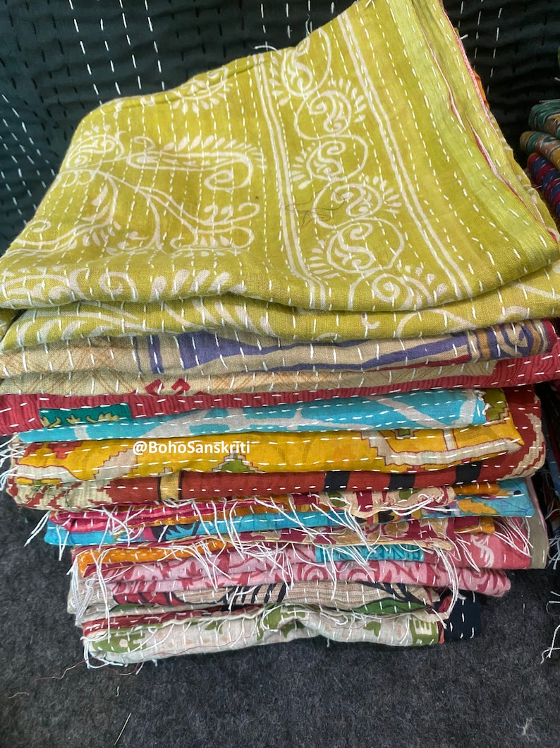 May include: A stack of colourful kantha quilts with intricate embroidery and stitching. The top quilt is a light green with white floral designs. The quilts are folded and stacked on top of each other. The text "@BohoSanskruti" is visible on the stack of quilts.