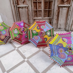 May include: Four colorful, patterned umbrellas with fringe trim. The umbrellas are made of fabric and have a variety of colors and designs, including red, blue, green, yellow, and pink. The umbrellas are arranged in a row on a white tiled floor.