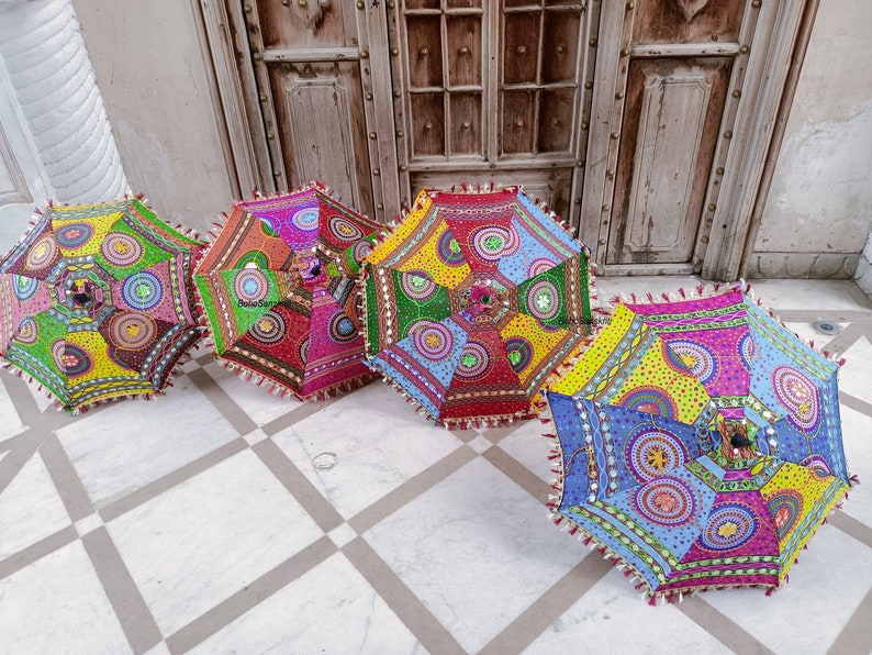 May include: Four colorful, patterned umbrellas with fringe trim. The umbrellas are made of fabric and have a variety of colors and designs, including red, blue, green, yellow, and pink. The umbrellas are arranged in a row on a white tiled floor.