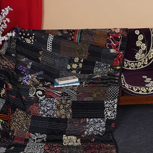 May include: Black and white kantha quilt with a patchwork design. The quilt is made of recycled saris and is hand-stitched. The quilt is folded and placed on a wooden bench with a purple cushion.