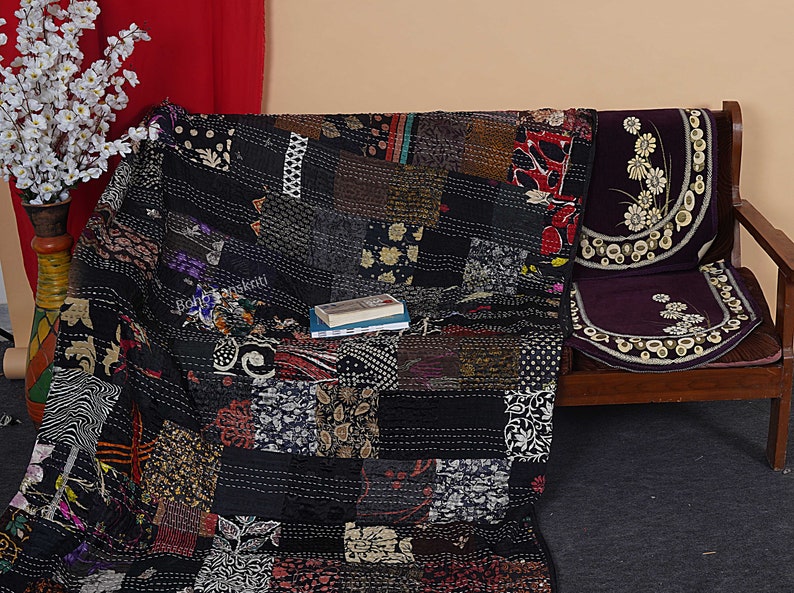 May include: Black and white kantha quilt with a patchwork design. The quilt is made of recycled saris and is hand-stitched. The quilt is folded and placed on a wooden bench with a purple cushion.