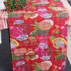 May include: Red kantha quilt table runner with a floral pattern. The runner features a variety of colourful flowers and leaves, including pink, orange, yellow, green, and purple. The runner is quilted with a fine stitch and has a fringed edge.