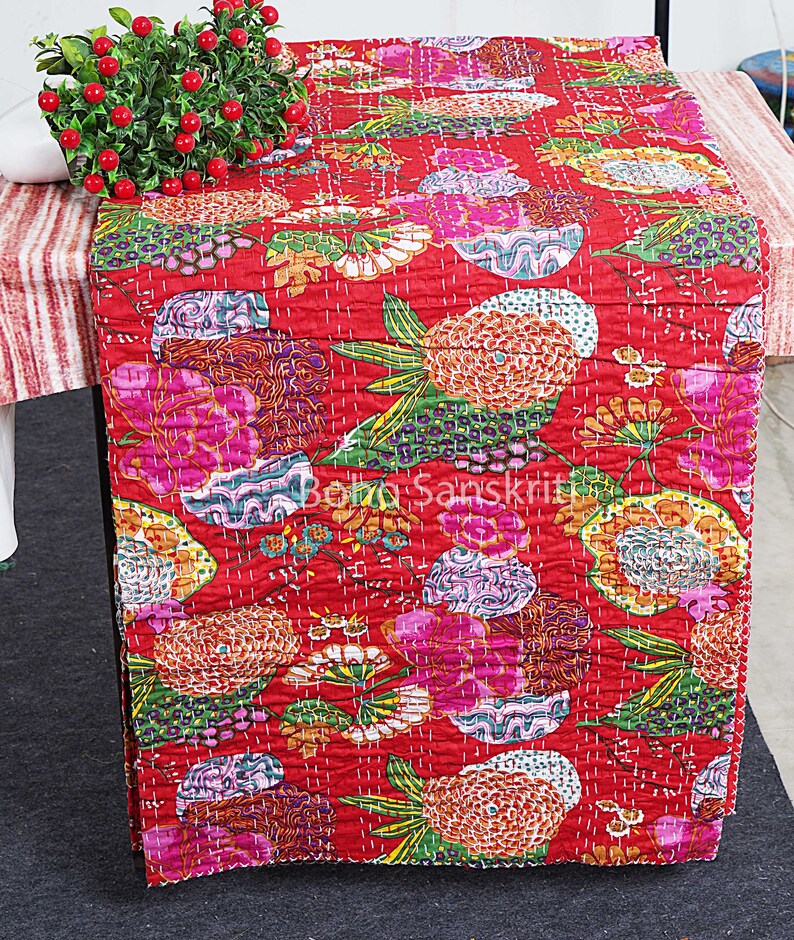 May include: Red kantha quilt table runner with a floral pattern. The runner features a variety of colourful flowers and leaves, including pink, orange, yellow, green, and purple. The runner is quilted with a fine stitch and has a fringed edge.