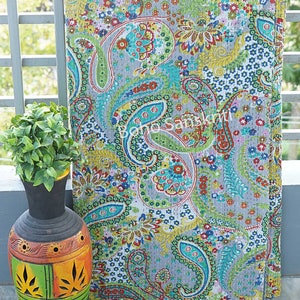 Op de afbeelding: Een grijze paisley-patroonquilt met een levendige mix van kleuren, waaronder groen, blauw, rood en geel. De quilt is gedrapeerd over een reling naast een decoratieve vaas met een plant.