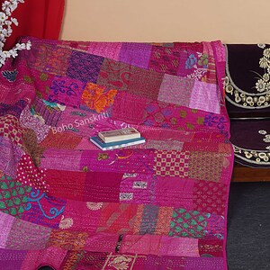 May include: A pink kantha quilt with a patchwork design. The quilt is made of various fabrics and is hand-stitched. The quilt is folded and placed on a wooden chair with a purple cushion. The text "boho sanskriti" is visible on the quilt.