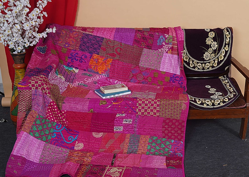 May include: A pink kantha quilt with a patchwork design. The quilt is made of various fabrics and is hand-stitched. The quilt is folded and placed on a wooden chair with a purple cushion. The text "boho sanskriti" is visible on the quilt.
