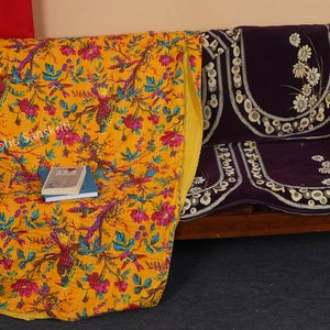 May include: A yellow kantha quilt with a floral and bird print, folded on a wooden bench with a purple cushion. The quilt has a yellow border and the cushion has white floral embroidery.