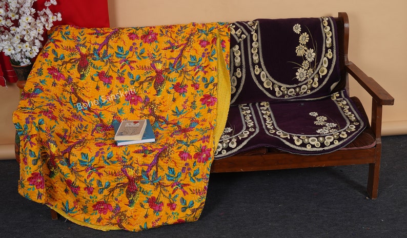 May include: A yellow kantha quilt with a floral and bird print, folded on a wooden bench with a purple cushion. The quilt has a yellow border and the cushion has white floral embroidery.