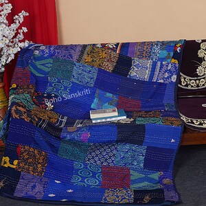 May include: A blue kantha quilt with a patchwork design featuring various colors and patterns. The quilt is made of recycled saris and is hand-stitched. The quilt is folded and placed on a wooden bench with a dark purple cushion. The text "Boho Sanskriti" is visible on the quilt.