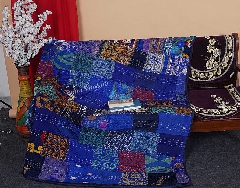 May include: A blue kantha quilt with a patchwork design featuring various colors and patterns. The quilt is made of recycled saris and is hand-stitched. The quilt is folded and placed on a wooden bench with a dark purple cushion. The text "Boho Sanskriti" is visible on the quilt.