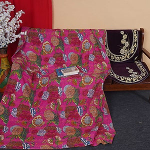 May include: A pink kantha quilt with a floral pattern. The quilt is draped over a wooden bench with a purple cushion. The quilt is made of cotton and has a hand-stitched design.