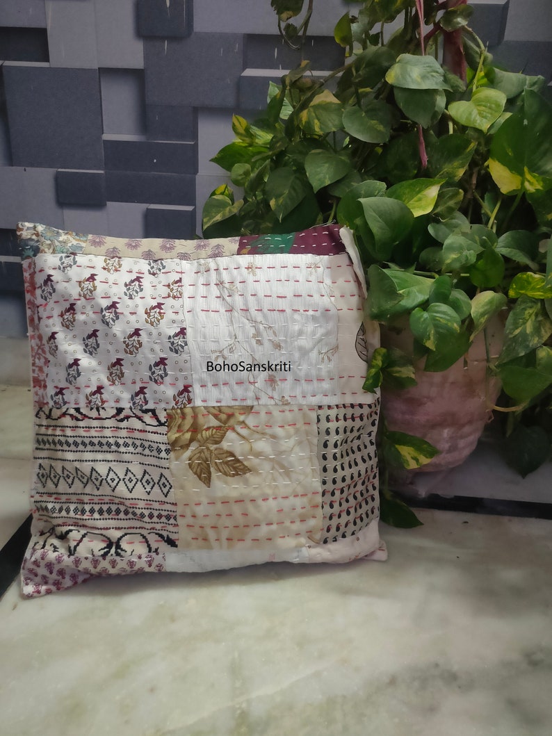 May include: Square patchwork pillow with a variety of patterned fabrics in cream, burgundy, and black. The pillow features visible stitching and the text "BohoSanskriti". A potted plant is in the background.