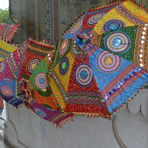 May include: A collection of colorful, patterned umbrellas with intricate embroidery and mirror embellishments. The umbrellas are hanging from a wall, creating a vibrant display of Indian craftsmanship.