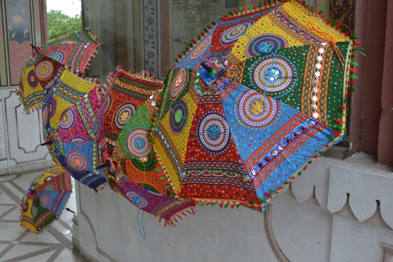 May include: A collection of colorful, patterned umbrellas with intricate embroidery and mirror embellishments. The umbrellas are hanging from a wall, creating a vibrant display of Indian craftsmanship.