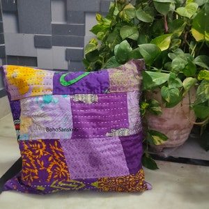 May include: A patchwork pillow in shades of purple, yellow, and orange. The pillow features a variety of fabric patterns and textures, with visible stitching. The text "BohoSanskriti" is printed on the pillow. The pillow is square and appears to be handmade.