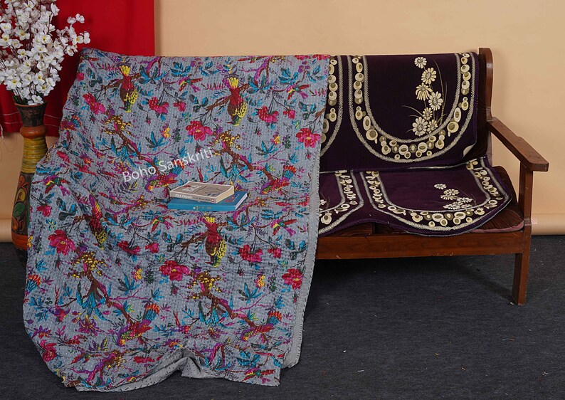May include: A gray and blue kantha quilt with a floral and bird pattern. The quilt is draped over a wooden bench with a purple cushion. The text "boho sanskriti" is visible on the quilt.