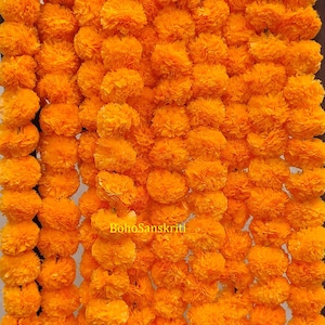 May include: A garland of bright orange marigold flowers, strung together in a long strand. The flowers are made of paper and are arranged in a repeating pattern.