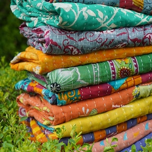 May include: A stack of colorful kantha quilts with floral patterns. The quilts are folded and stacked on top of each other, creating a vibrant and textured display. The text "boho sanskriti" is visible on one of the quilts.