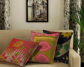 May include: Four decorative throw pillows on a beige sofa. The pillows feature various patterns, including floral and leaf designs in red, green, and black. A framed abstract artwork hangs above the sofa.