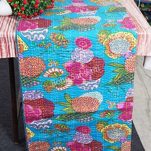 May include: A turquoise blue table runner with a floral pattern. The flowers are in shades of pink, orange, yellow, and green. The runner is quilted and has a subtle sheen.