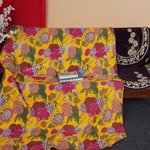 May include: A yellow kantha quilt with a floral pattern in shades of pink, green, and yellow. The quilt is draped over a wooden bench with a purple cushion.