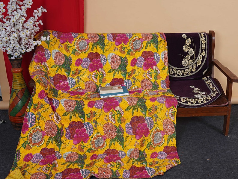 May include: A yellow kantha quilt with a floral pattern in shades of pink, green, and yellow. The quilt is draped over a wooden bench with a purple cushion.
