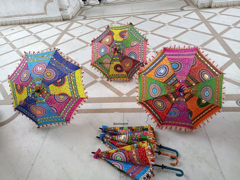 May include: Three colorful, patterned umbrellas with fringe trim and sequins. The umbrellas are laid out on a white tiled floor.
