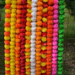 May include: A set of 10 garlands made of artificial marigold flowers. The garlands are in various colours, including yellow, orange, red, white, pink, and green. The garlands are hanging on a wire fence.