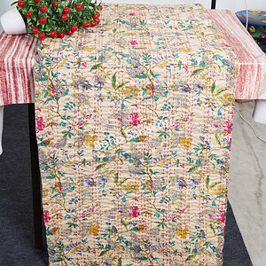 以下が含まれることがあります： ベージュのテーブルランナーで、ピンク、ブルー、グリーン、イエローの花柄が施されています。ランナーはキルト加工されており、上品な光沢があります。