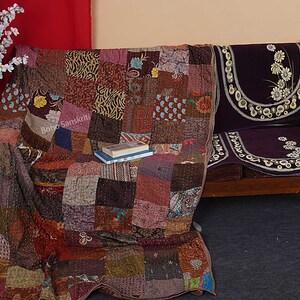 May include: A patchwork quilt made of various fabrics in shades of brown, pink, and blue. The quilt is draped over a wooden couch with a purple velvet cushion. The couch is decorated with a floral pattern.