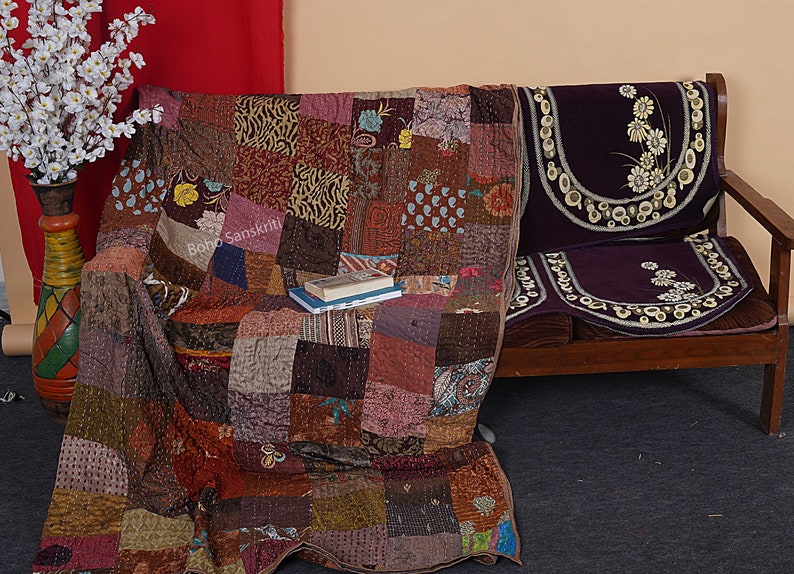 May include: A patchwork quilt made of various fabrics in shades of brown, pink, and blue. The quilt is draped over a wooden couch with a purple velvet cushion. The couch is decorated with a floral pattern.