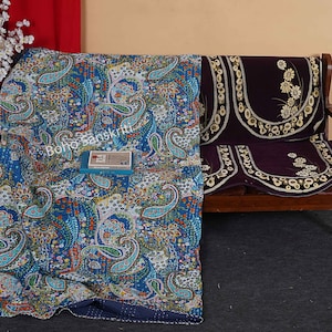 May include: A blue and white paisley patterned kantha quilt with a dark blue border. The quilt is folded and placed on a wooden bench with a purple and gold floral patterned cushion.