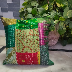 May include: Square patchwork pillow with a variety of green, red, and pink fabric pieces. The pillow has white stitching throughout and the text "BohoSanskriti" is visible. The pillow is placed in front of a plant.