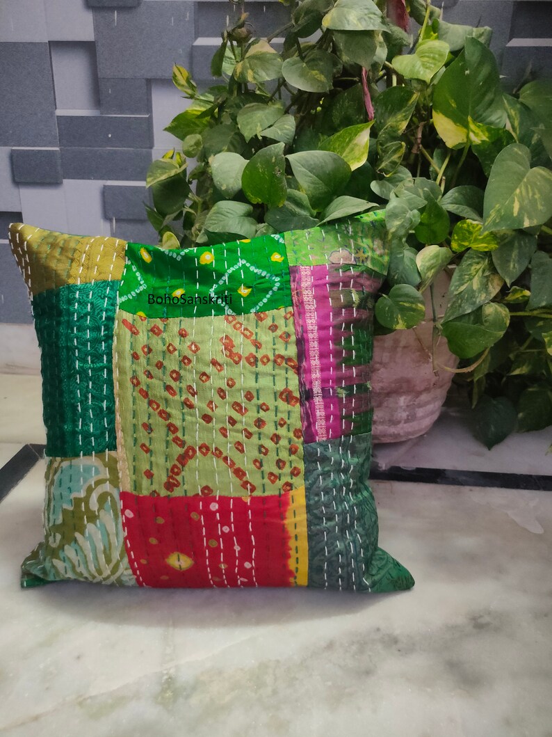 May include: Square patchwork pillow with a variety of green, red, and pink fabric pieces. The pillow has white stitching throughout and the text "BohoSanskriti" is visible. The pillow is placed in front of a plant.