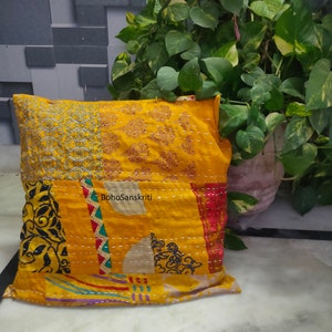 May include: A decorative yellow pillow with patchwork design. The pillow features embroidered patterns and the text "BohoSanskriti". The pillow is placed on a white surface with a green plant in the background.