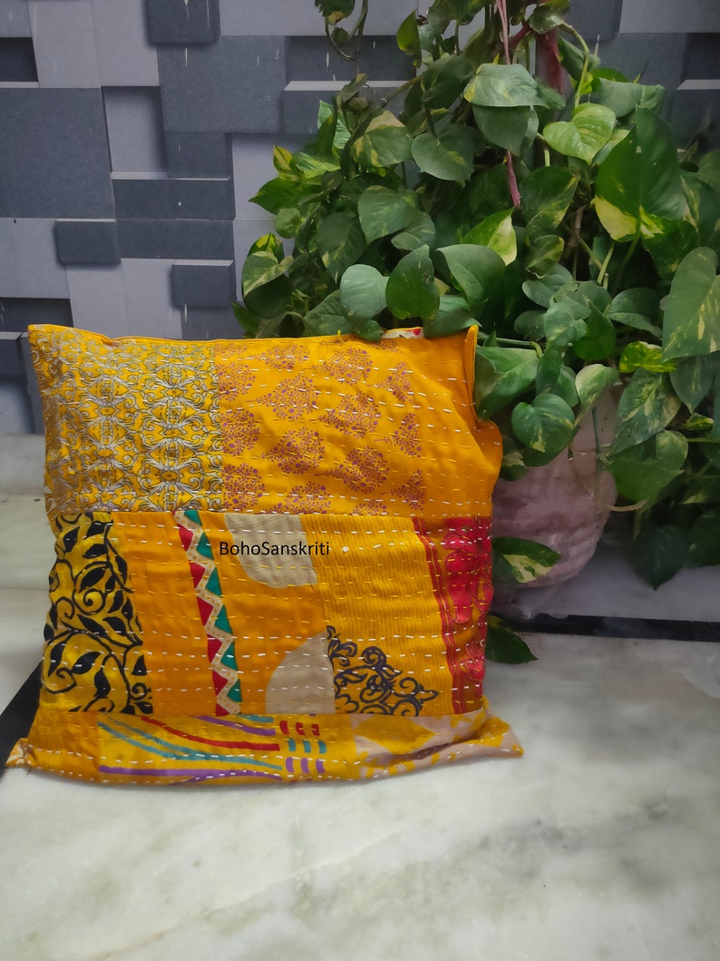 May include: A decorative yellow pillow with patchwork design. The pillow features embroidered patterns and the text "BohoSanskriti". The pillow is placed on a white surface with a green plant in the background.