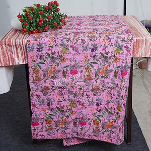 以下が含まれることがあります： 白い縁取りのあるピンクの花柄カンタキルト。キルトは綿製で、手縫いのデザインが施されています。キルトは折り畳んでテーブルに飾られています。