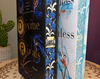 May include: Two paperback books with floral designs on the covers. The book on the left is titled "Divine Rivals" by Rebecca Ross. The book on the right is titled "The Endless" by Rebecca Ross.