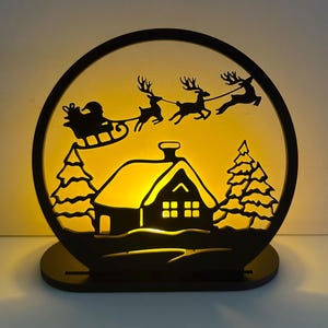 Op de afbeelding: Een zwarte, cirkelvormige kerstversiering met een silhouet van de slee van de kerstman getrokken door rendieren over een huis en bomen. De scène wordt verlicht met een warm geel licht, wat een feestelijke sfeer creëert. De decoratie staat op een zwarte basis.