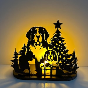 Puede incluir: Silueta negra de dos perros de montaña berneses con un árbol de Navidad y una estrella. Los perros están colocados frente a una luz amarilla brillante, con una caja de regalo frente al perro más pequeño. El diseño está sobre una base negra.