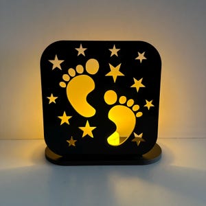 Puede incluir: Una luz de noche cuadrada negra con un diseño recortado con huellas de bebé y estrellas. Las huellas y las estrellas están iluminadas con un brillo amarillo cálido, creando una luz suave. La luz de noche se encuentra sobre una base redonda pequeña.