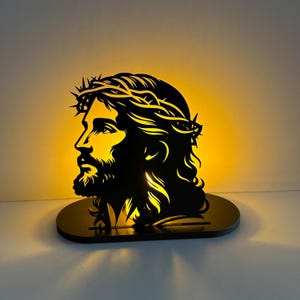Puede incluir: Una silueta negra del perfil de Jesús, con una corona de espinas, iluminada desde atrás con una cálida luz amarilla. La imagen es una pieza decorativa, probablemente una lámpara o un adorno de pared, con un diseño recortado detallado.