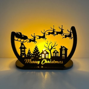 Könnte beinhalten: Schwarze Weihnachtsdekoration aus Holz mit einer Silhouette von Santa's Schlitten, der von Rentieren über eine Stadt gezogen wird. Die Worte "Merry Christmas" sind in die Basis geschnitten. Der Hintergrund leuchtet mit warmem gelbem Licht.