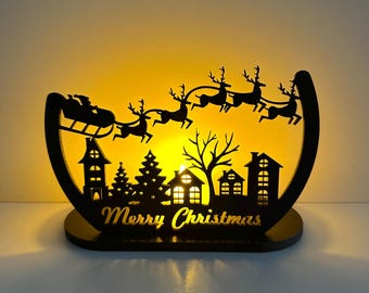 Könnte beinhalten: Schwarze Weihnachtsdekoration aus Holz mit einer Silhouette von Santa's Schlitten, der von Rentieren über eine Stadt gezogen wird. Die Worte "Merry Christmas" sind in die Basis geschnitten. Der Hintergrund leuchtet mit warmem gelbem Licht.