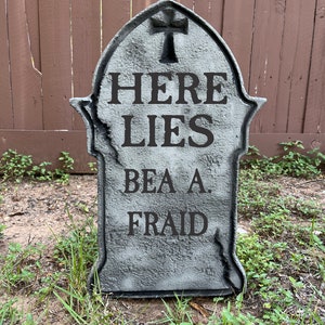 May include: A gray tombstone with the text "HERE LIES BEA A FRAID" on it. The tombstone is in a grassy area with a brown wooden fence in the background.