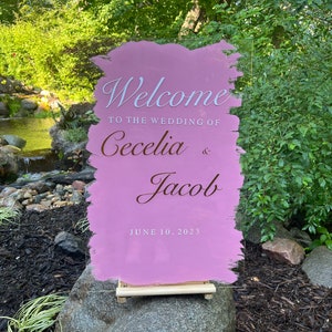 May include: Pink acrylic wedding welcome sign with white brushstrokes. The sign reads "Welcome to the wedding of Cecelia & Jacob June 10, 2023".