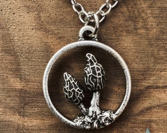 May include: Silver necklace with a round pendant featuring a detailed depiction of two morel mushrooms. The pendant is on a silver chain.
