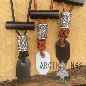 Może przedstawiać: Trzy skórzane naszyjniki z czarnymi drewnianymi koralikami i srebrnymi zawieszkami. Zawieszki przedstawiają wzór węzła i symbol runy. Naszyjniki mają czarne i białe drewniane zawieszki.