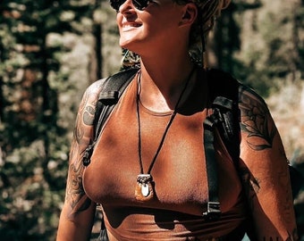 May include: A rust-colored sleeveless crop top paired with black athletic shorts and a plaid flannel shirt tied around the waist. The outfit includes a black backpack, a dark pendant necklace, and black sunglasses, worn while hiking in a forest.
